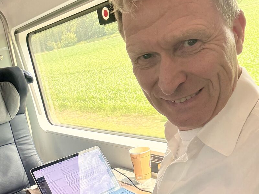 Dr. Christoph Wesselmann im Zug am Fensterplatz mit aufgeklapptem Laptop und Kaffee — während fünf KI-Agenten parallel arbeiten, bleibt Zeit für eine Pause